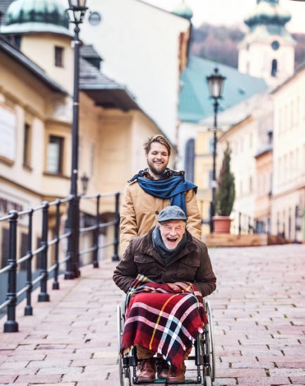 Father in wheelchair and young son on a walk, laughing. A carer assisting disabled senior man.